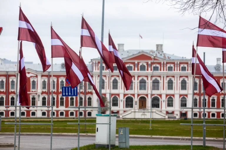 Plusieurs drapeaux lettons flottant devant un palais historique rouge et blanc par temps gris.