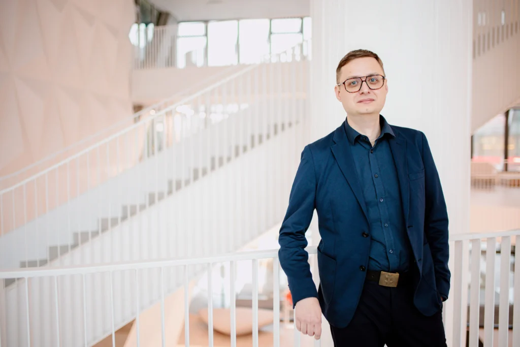 Vīrietis brillēs un tumši zilā uzvalkā stāv pie baltām kāpnēm mūsdienīgā, gaišā telpā.