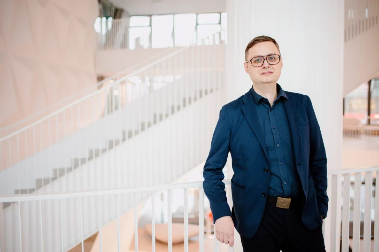 Vīrietis brillēs un tumši zilā uzvalkā stāv pie baltām kāpnēm mūsdienīgā, gaišā telpā.