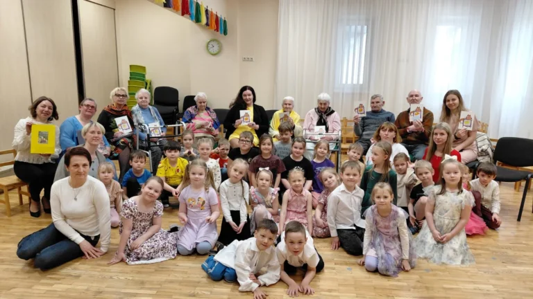 Liela bērnu un senioru grupa telpā uz koka grīdas, turot rokās krāsainas kartītes.