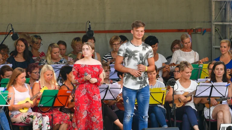 Jauni mākslinieki uzstājas uz skatuves brīvdabas koncertā, ko papildina plašs instrumentālais pavadījums.