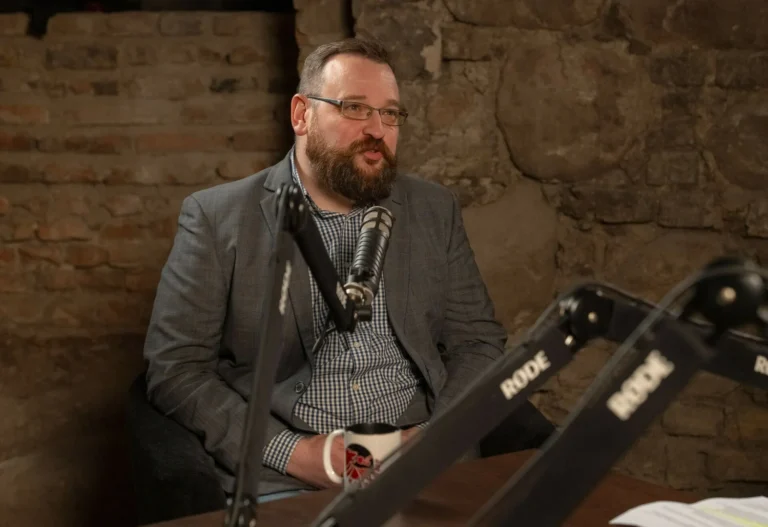 Bārdains vīrietis brillēs un žaketē sēž pie mikrofona podkāsta studijā uz mūra sienas fona.