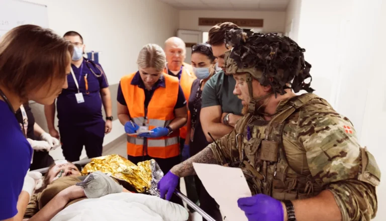 Karavīrs maskēšanās tērpā sarunājas ar medicīnas darbiniekiem pie pacienta uz nestuvēm slimnīcas telpās.