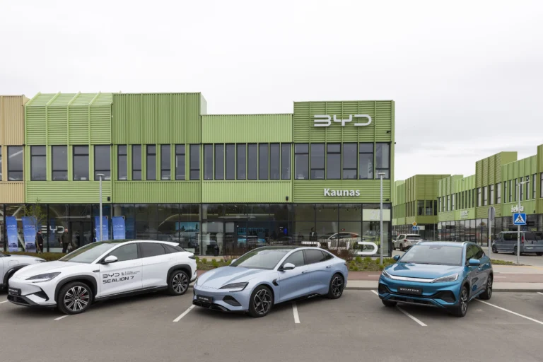 Trīs BYD elektromobiļi novietoti stāvvietā pie modernas zaļas krāsas automašīnu salona ēkas Kauņā.