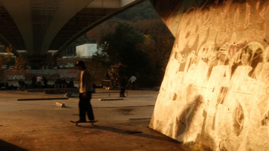 Skeitbordists zem tilta saulrietā pie grafiti sienas, simbolizējot pilsētas jauniešu kultūru un aktīvo dzīvesveidu.