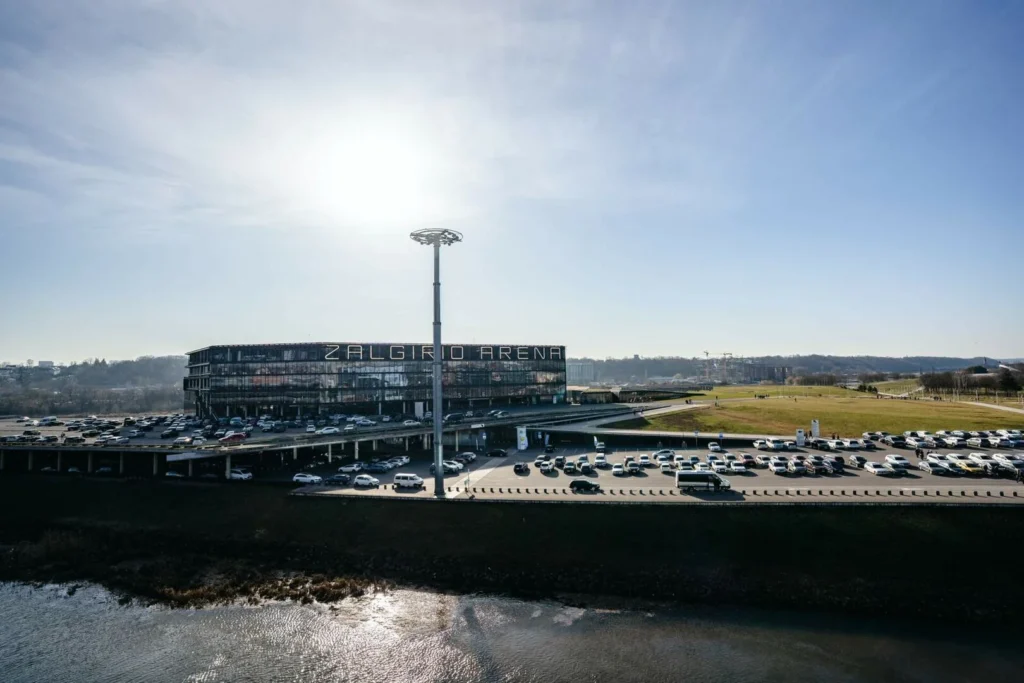 Mūsdienīgā Žalgirio Arena ēka pie upes ar plašu autostāvvietu saulainā dienā.