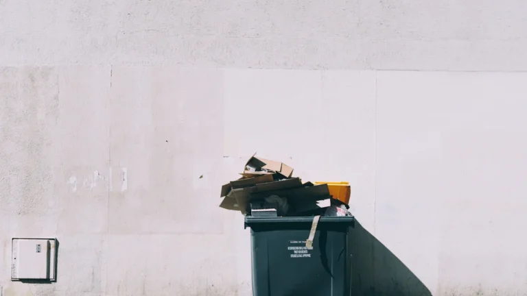 Melns plastmasas atkritumu konteiners ar riteņiem novietots pie gaišas ēkas sienas.