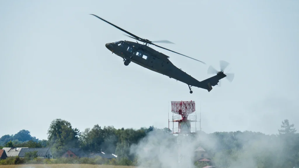 Melns militārais helikopters pacelšanās laikā virs dūmu mākoņa un radara torņa gaišā dienas laikā.