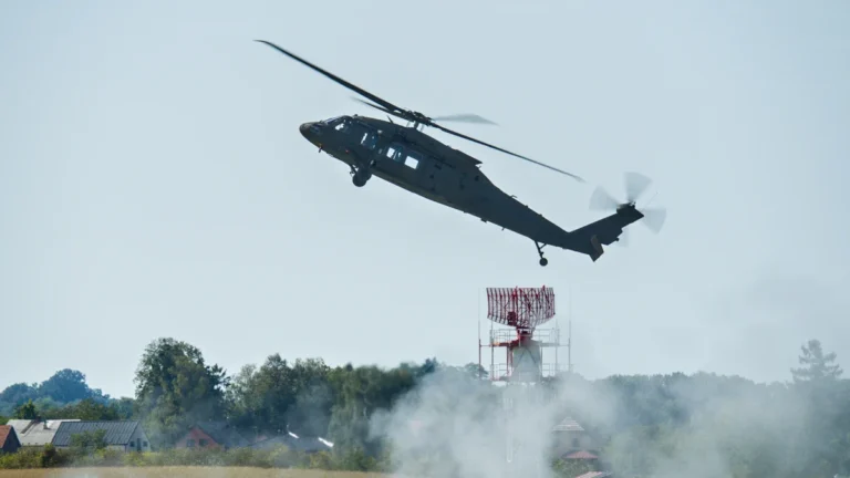 Melns militārais helikopters pacelšanās laikā virs dūmu mākoņa un radara torņa gaišā dienas laikā.