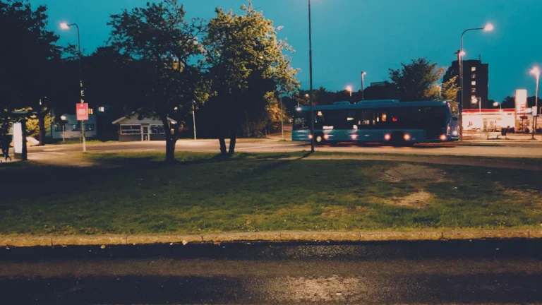 Zilas krāsas pilsētas autobuss brauc pa ielu vēlā vasaras vakarā gar zaļojošiem kokiem.