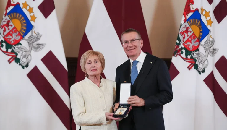 Latvijas prezidents Edgars Rinkēvičs un sieviete gaišā tērpā kopā tur atvērtu apbalvojuma kārbu.