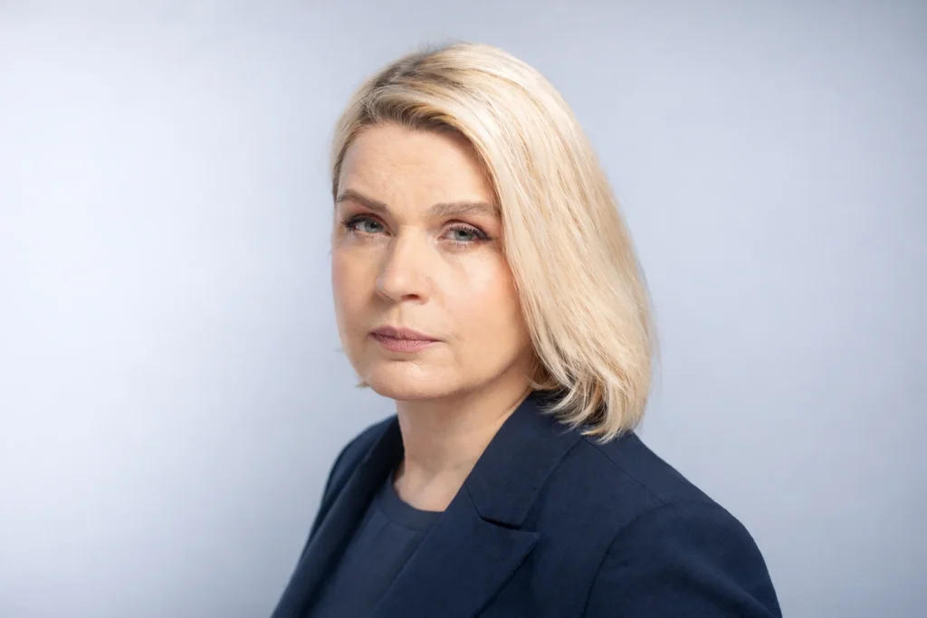 Blonda sieviete tumši zilā žaketē ar nopietnu sejas izteiksmi uz gaiši pelēka fona.