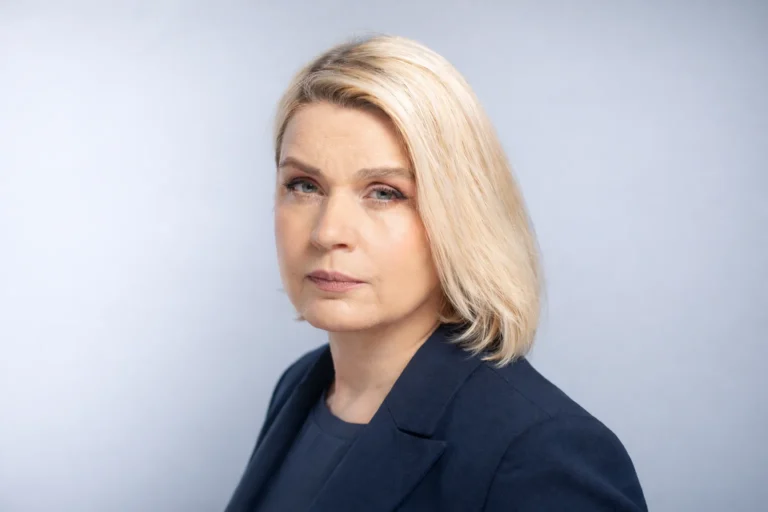 Blonda sieviete tumši zilā žaketē ar nopietnu sejas izteiksmi uz gaiši pelēka fona.