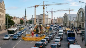 Rīgas iela ar intensīvu satiksmi, būvniecības celtņiem, ekskavatoriem un vēsturiskām ēkām pilsētas centrā.