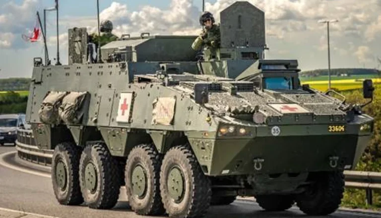 Liela zaļa astoņriteņu militārā medicīniskā transportmašīna ar sarkaniem krustiem un diviem karavīriem uz šosejas.