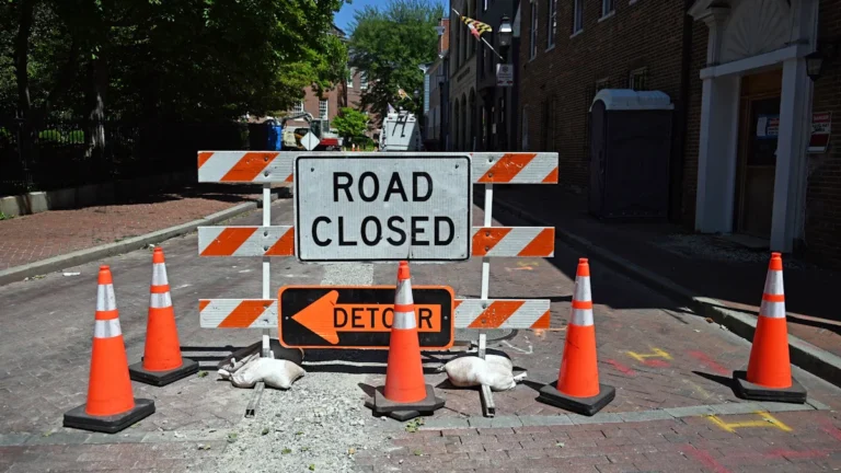 Ceļa zīme 'Road Closed' un barjeras ar oranžiem konusiem uz ielas.