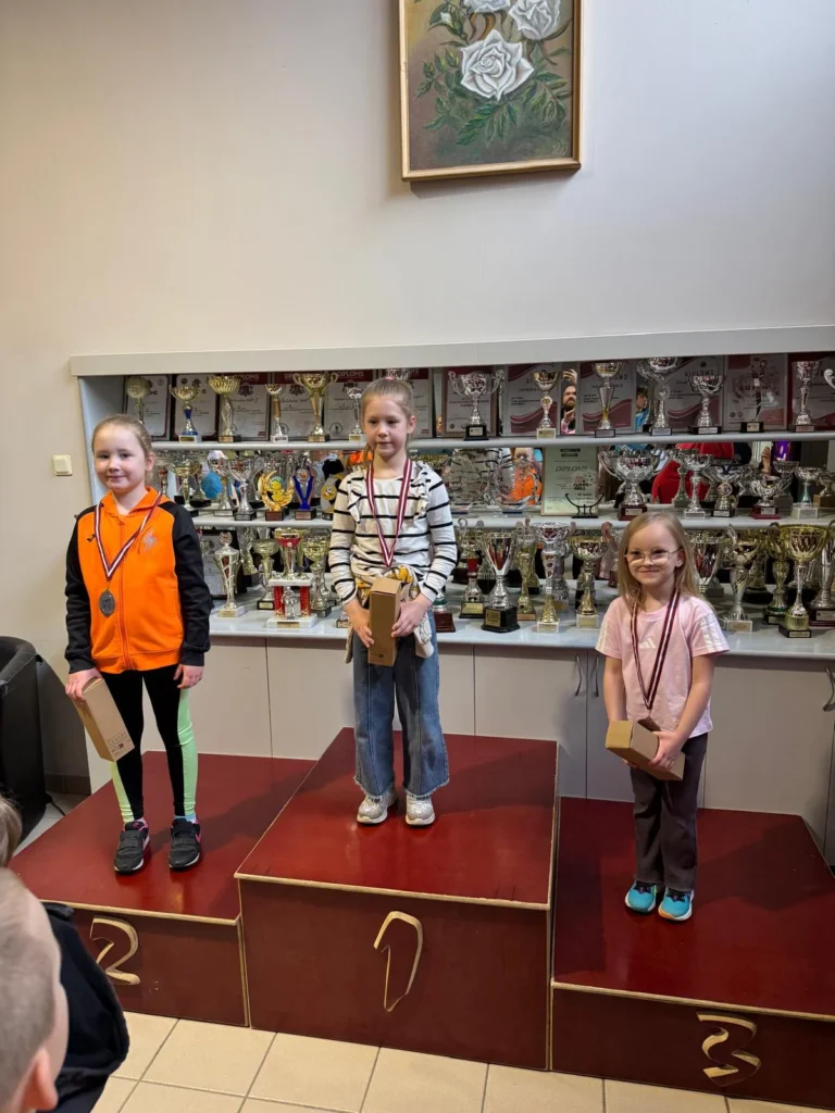 Trīs jaunas meitenes stāv uz goda pjedestāla ar medaļām kaklā un dāvanām rokās.