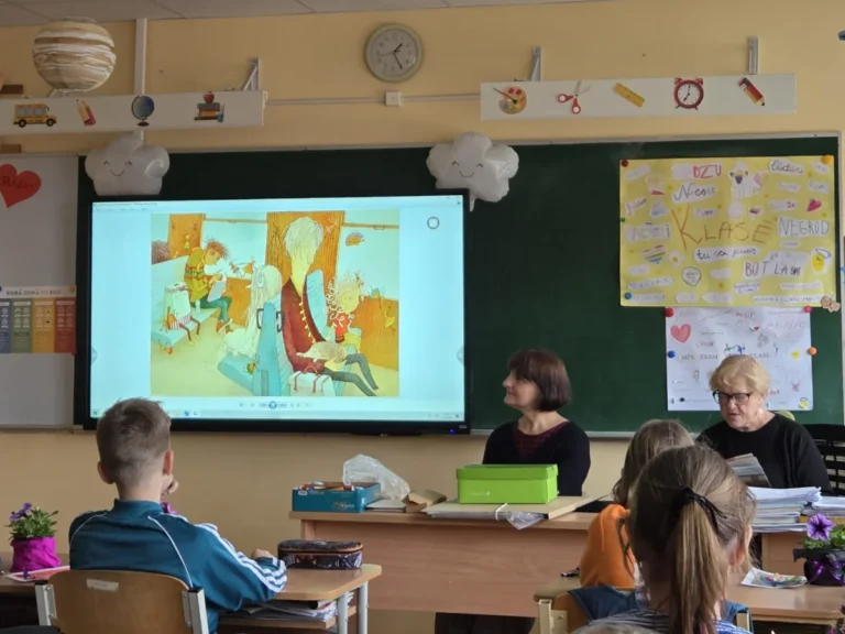 Skolotājas klases priekšā pie interaktīvā ekrāna ar ilustrāciju, skolēni sēž solos un vēro.