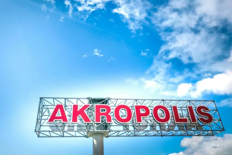 Liela sarkana izkārtne ar uzrakstu 'AKROPOLIS' uz metāla konstrukcijas pret gaiši zilām debesīm ar mākoņiem.