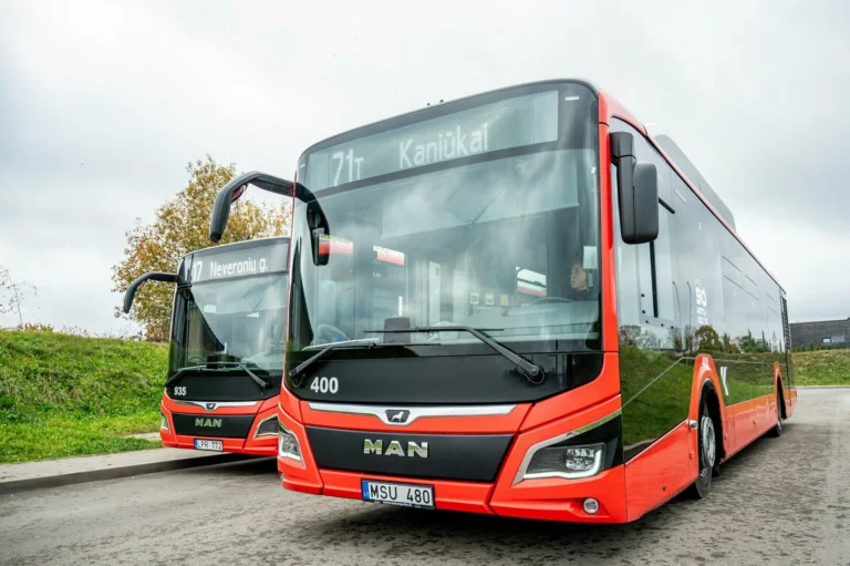 Divi mūsdienīgi sarkani un melni MAN pilsētas autobusi stāv uz ceļa zem mākoņainām debesīm.