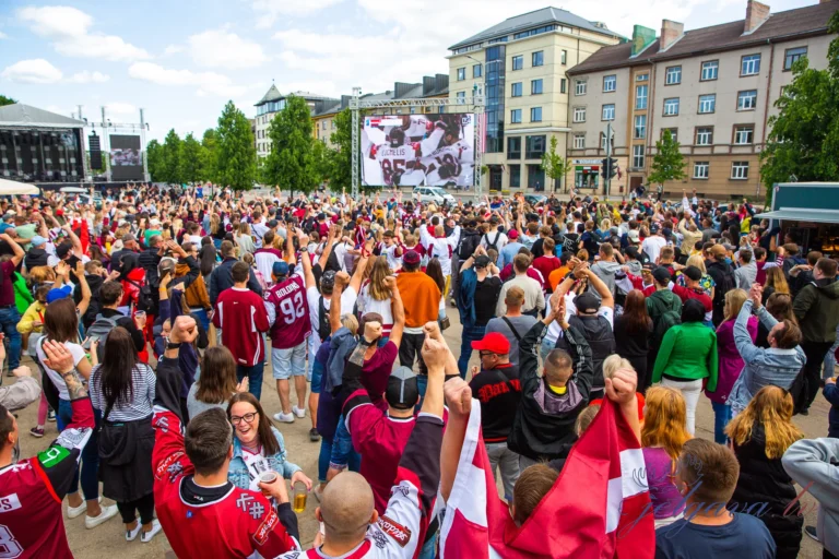 Liels pūlis cilvēku Latvijas hokeja krekliem vēro spēli uz liela ekrāna pilsētas laukumā.
