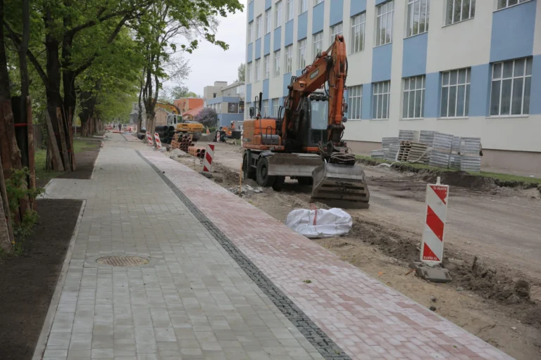 Oranžs ekskavators atrodas uz ielas remontdarbu joslas blakus tikko ieklātam bruģim un lielai ēkai.