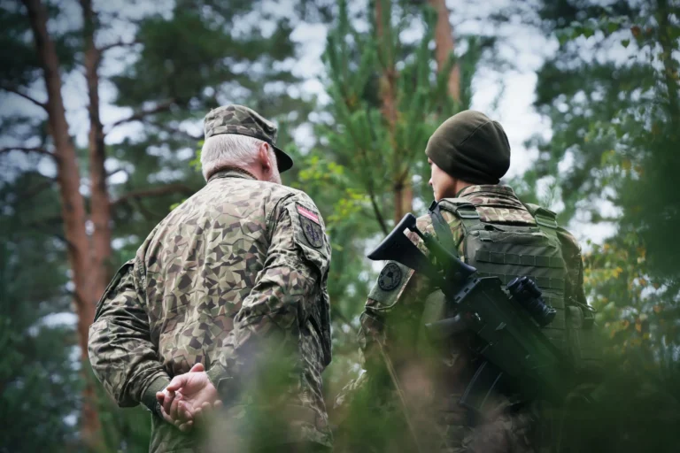 Divi karavīri Latvijas armijas formas tērpos stāv mežā, vienam uz muguras ir triecienšautene.