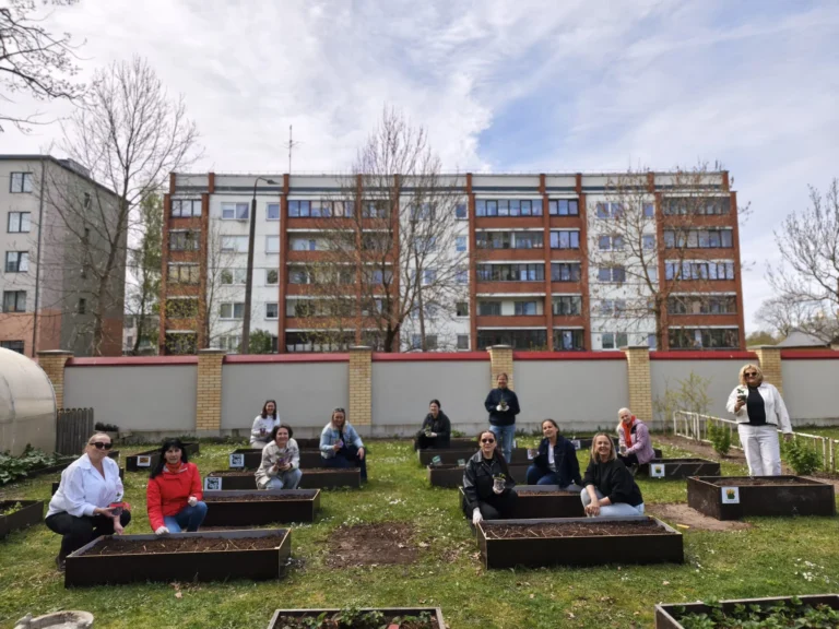 Sieviešu grupa pozē ar stādiem pie paceltām dobēm pilsētas pagalmā uz daudzdzīvokļu mājas fona.