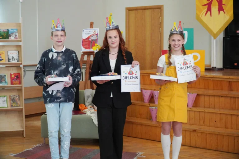 Trīs bērni ar papīra kroņiem galvā stāv telpā, turot rokās diplomus un grāmatas.