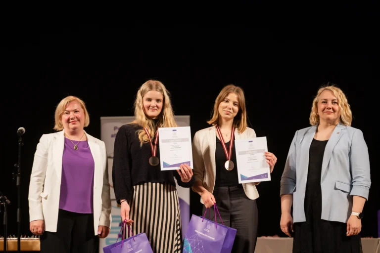 Divas godalgotas jaunietes ar diplomiem un medaļām stāv kopā ar divām sievietēm uz tumša fona.