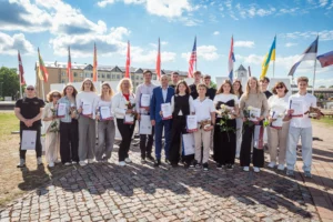 Grupa jauniešu un pieaugušo pozē ar diplomiem un ziediem uz dažādu valstu karogu fona.