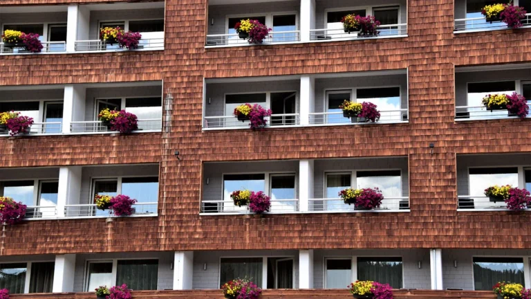 Daudzstāvu mājas fasāde ar daudziem balkoniem, kuri izrotāti ar košiem ziediem pilsētas vidē.
