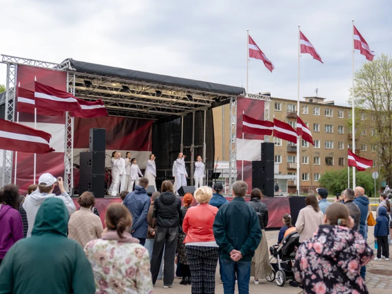 Jauniešu vokālā grupa baltos tērpos uzstājas uz brīvdabas skatuves, ko ieskauj daudzi Latvijas karogi.