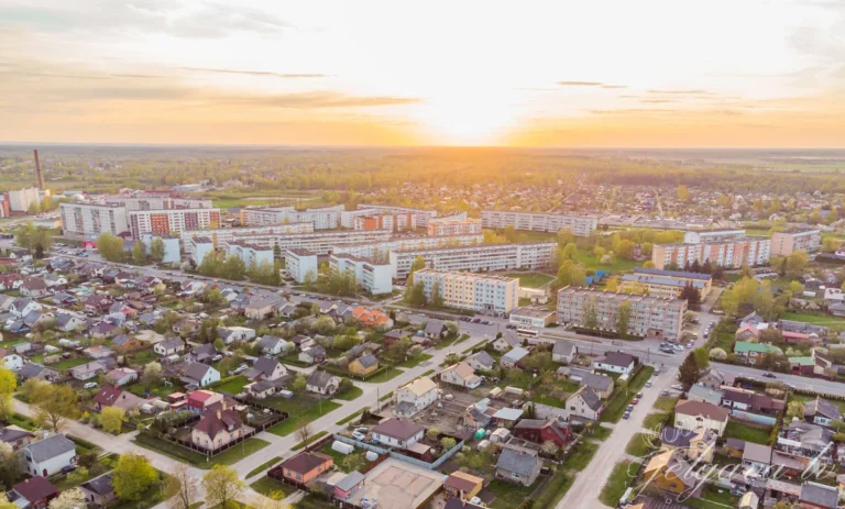 Aerofotoattēls ar Jelgavas pilsētu saulrietā, attēlojot privātmājas un daudzdzīvokļu ēkas siltā saules gaismā.