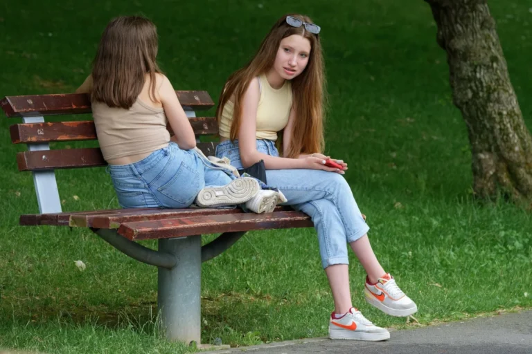 Divas jaunas meitenes ar gariem matiem un džinsiem sēž uz koka soliņa zaļā parkā.