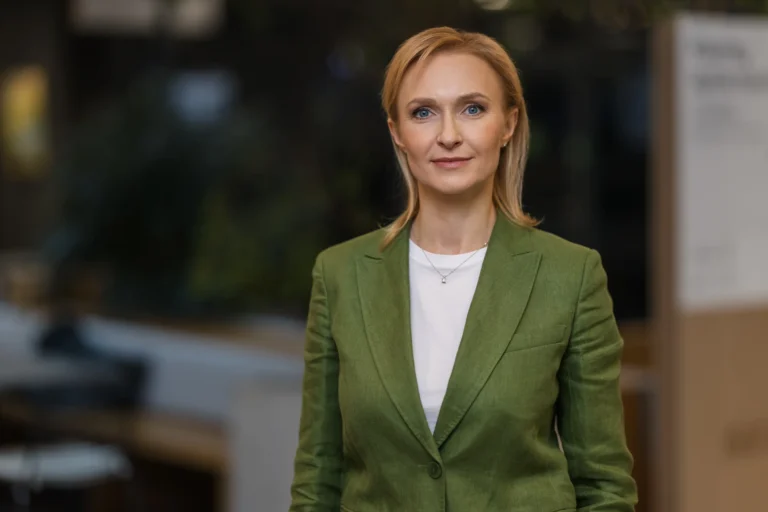Blondīne ar zilām acīm zaļā žaketē un baltā kreklā uz izpludināta biroja fona.