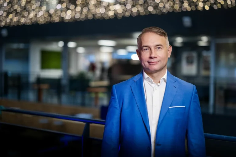Vīrietis spilgti zilā uzvalkā un baltā kreklā stāv modernā telpā uz izplūduša gaismu fona.