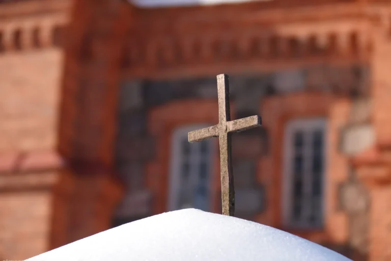 Neliels, teksturēts krusts iestiprināts sniega kupenā uz izplūduša sarkanu ķieģeļu baznīcas fasādes fona.