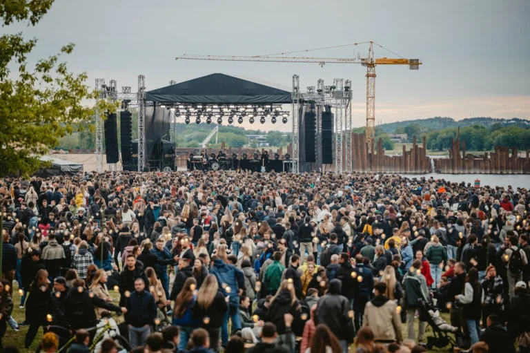Liels cilvēku pūlis stāv pie brīvdabas koncertu skatuves upes krastā saulrieta laikā.