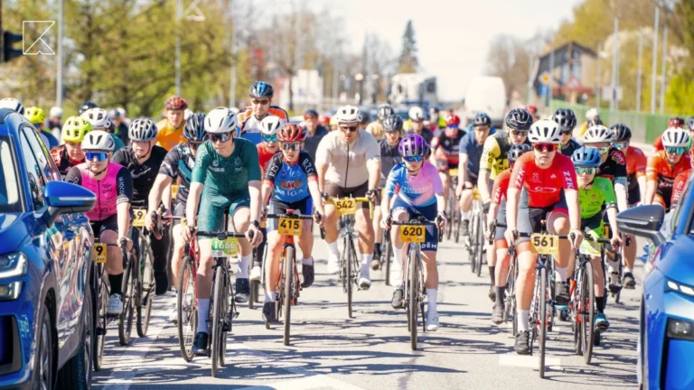 Liela velosipēdistu grupa krāsainos sporta tērpos brauc pa saulainu pilsētas ielu sacensību laikā.