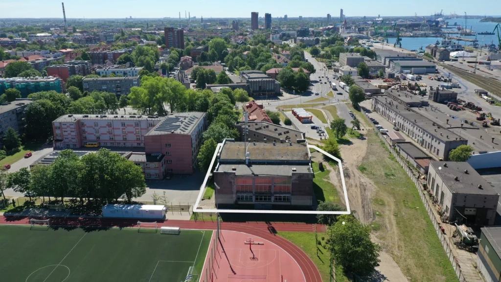 Pilsētas skats no putna lidojuma ar baltā rāmī iezīmētu ēku blakus sporta laukumam un ostu.