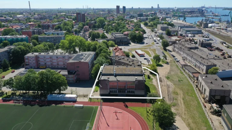 Pilsētas skats no putna lidojuma ar baltā rāmī iezīmētu ēku blakus sporta laukumam un ostu.