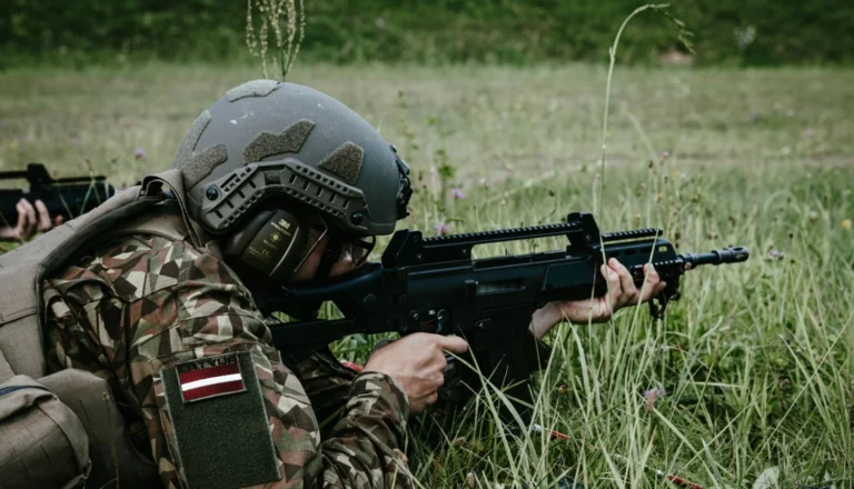 Karavīrs maskēšanās tērpā ar Latvijas karoga emblēmu tēmē ar triecienšauteni zālājā militāro mācību laikā.