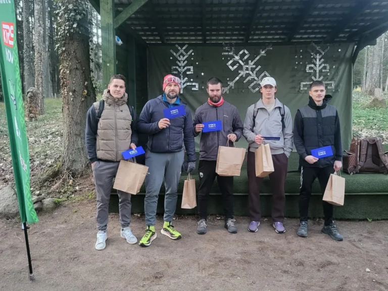 Pieci vīrieši sportiskā apģērbā stāv pie koka nojumes, turot rokās zilas dāvanu kartes un maisiņus.