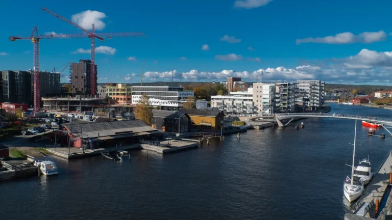 Pilsētas skats ar jaunceltnēm un celtniecības krāniem pie ūdens kanāla, simbolizējot pilsētvides attīstību.