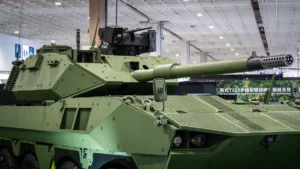 Mūsdienīgs bruņutransportieris ar lielgabalu aizsardzības izstādē, kas simbolizē fokusu uz jaunajām militārajām tehnoloģijām.