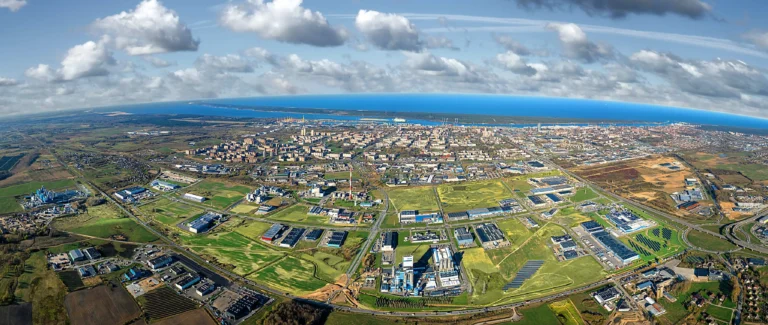 Pilsētas panorāma no augšas ar rūpniecības ēkām, zaļiem laukiem un zilo jūru pie apvāršņa.