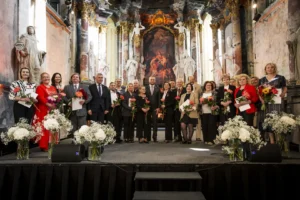 Cilvēku grupa ar ziediem un apliecībām rokās stāv uz skatuves greznā baznīcas interjerā.