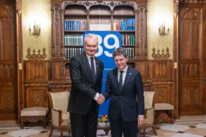 Divi vīrieši uzvalkos spiež rokas greznā telpā pie grāmatplaukta un zila B9 banera.
