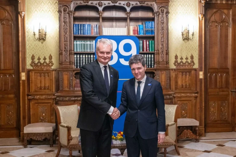 Divi vīrieši uzvalkos spiež rokas greznā telpā pie grāmatplaukta un zila B9 banera.